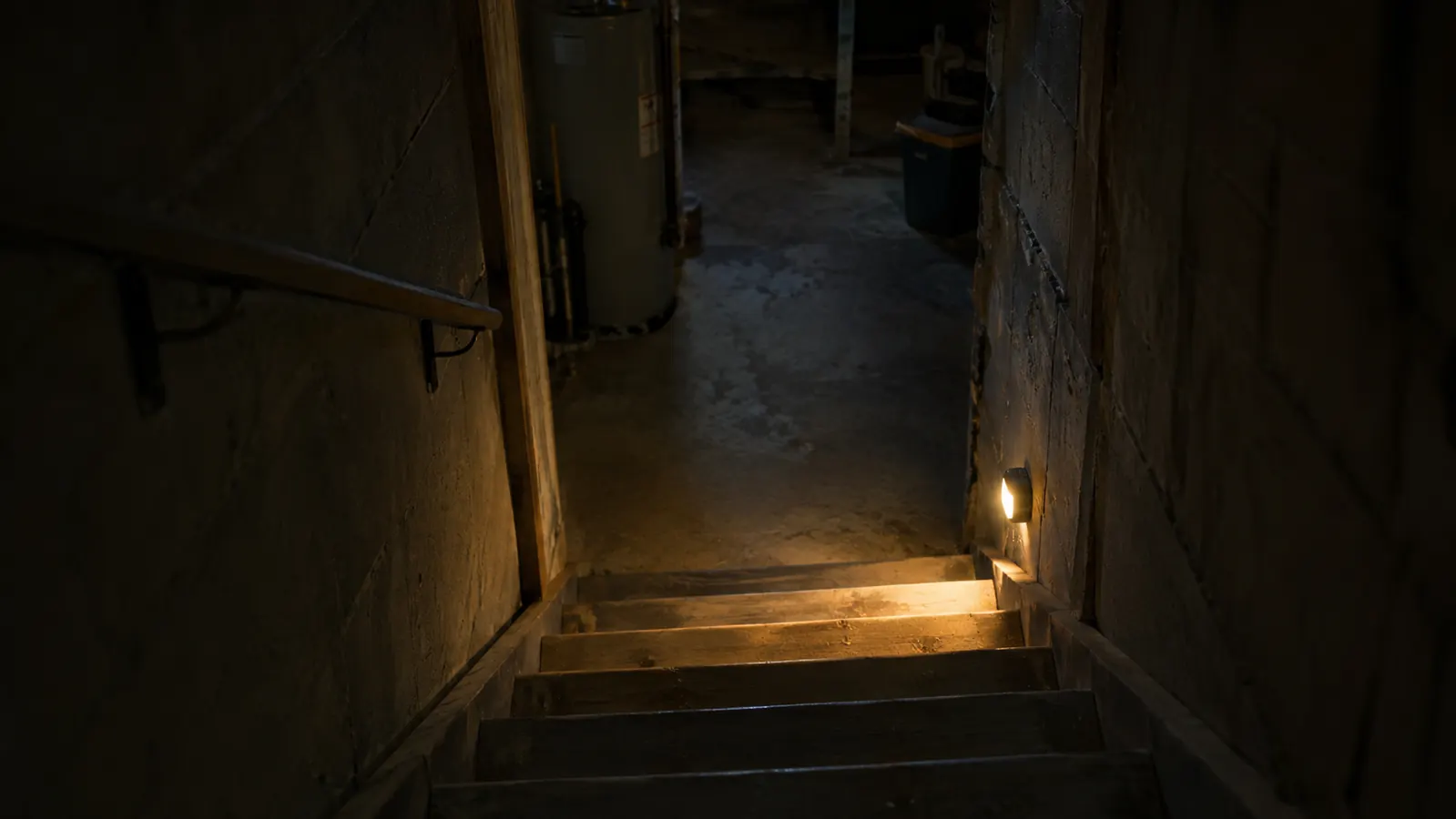A dark basement stairway at night lit by a small motion sensor light mounted near the bottom step, casting a pool of light on the stairs.